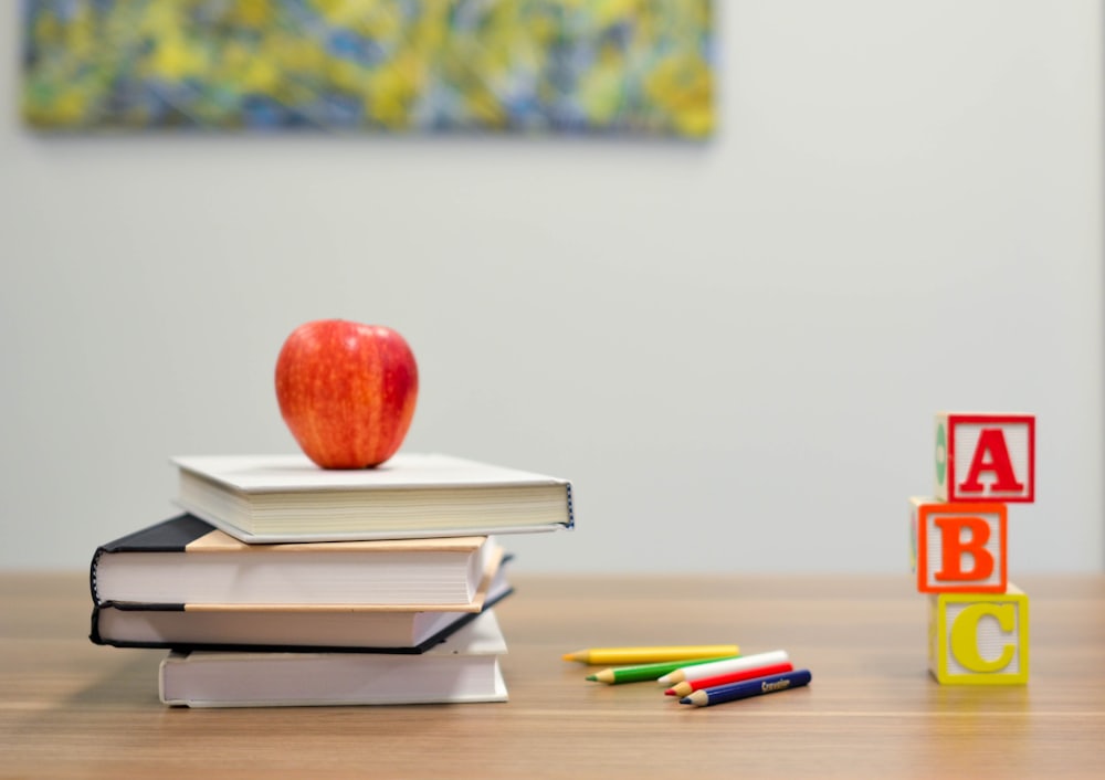 A neat stack of textbooks with a red apple and alphabet blocks resting on a classroom desk.